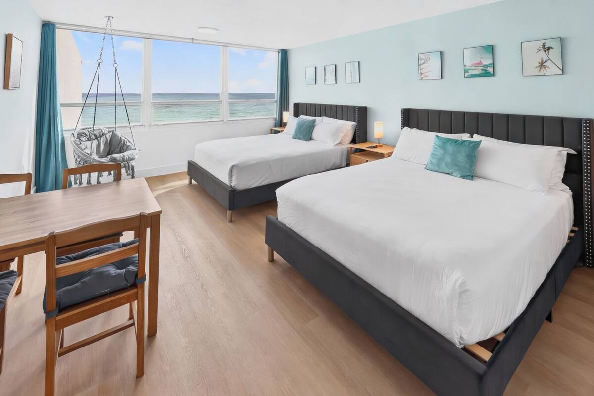 A hotel room with two beds, a desk, chairs, and a view of the ocean.