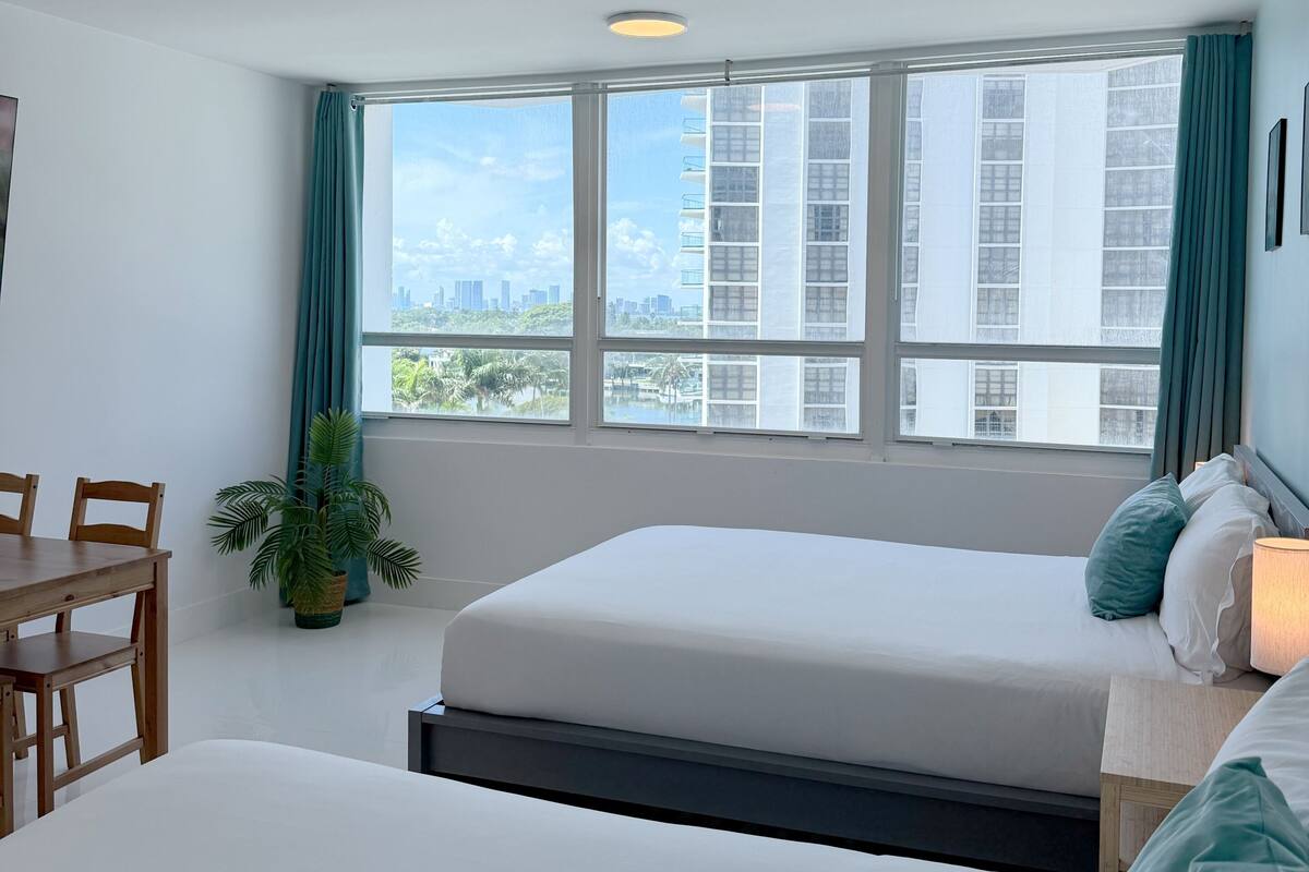 A hotel room with a bed, a desk, a chair, a potted plant, and a view of the cityscape through large windows.