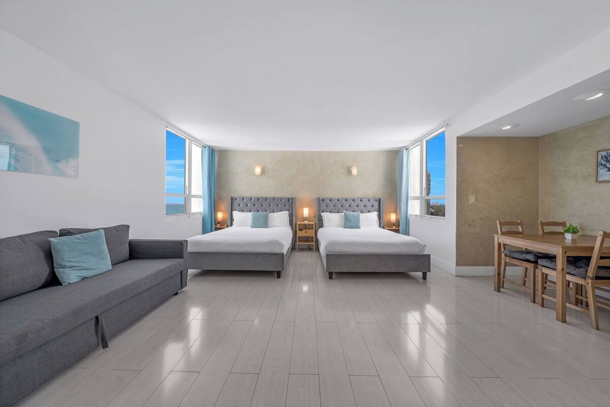 A modern hotel room with two beds, a sofa, a dining table, and large windows.