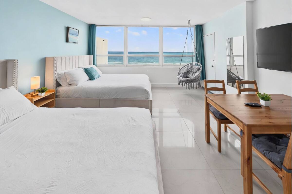 A hotel room with two beds, a dining table, a TV, and a view of the ocean.