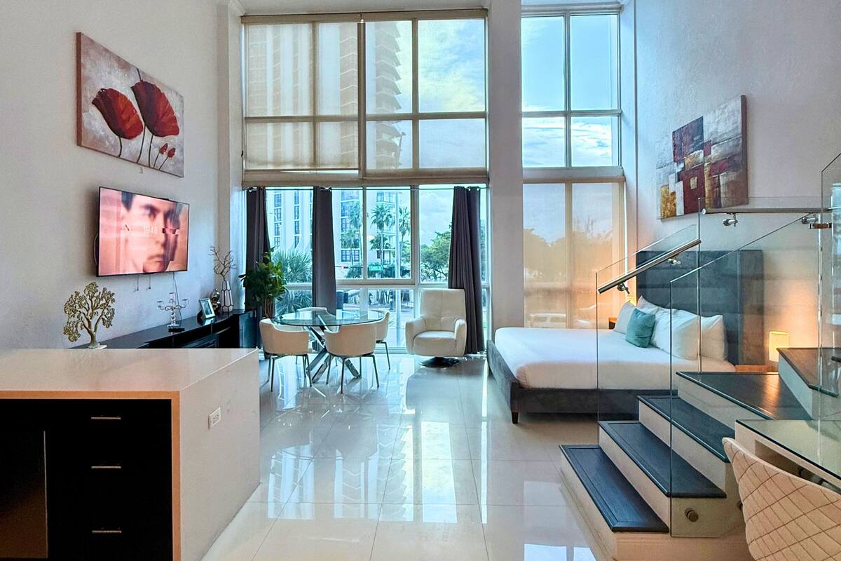 A modern hotel room with a large window, a dining area, and a staircase.