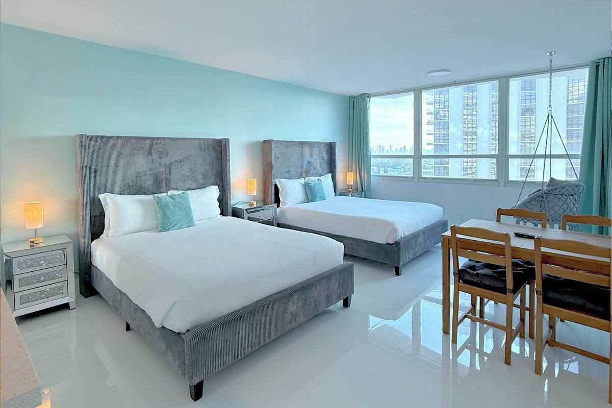 A hotel room with two beds, a dining table, chairs, a desk, and a window with a city view.