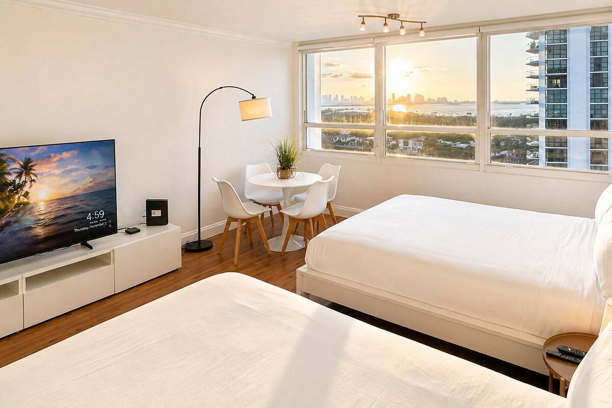 A hotel room with two beds, a desk with chairs, a TV, and a large window with a city view.