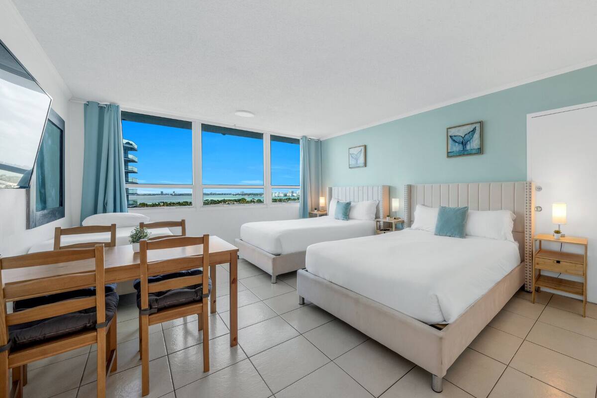 A hotel room with two beds, a dining table, and a view of the city.