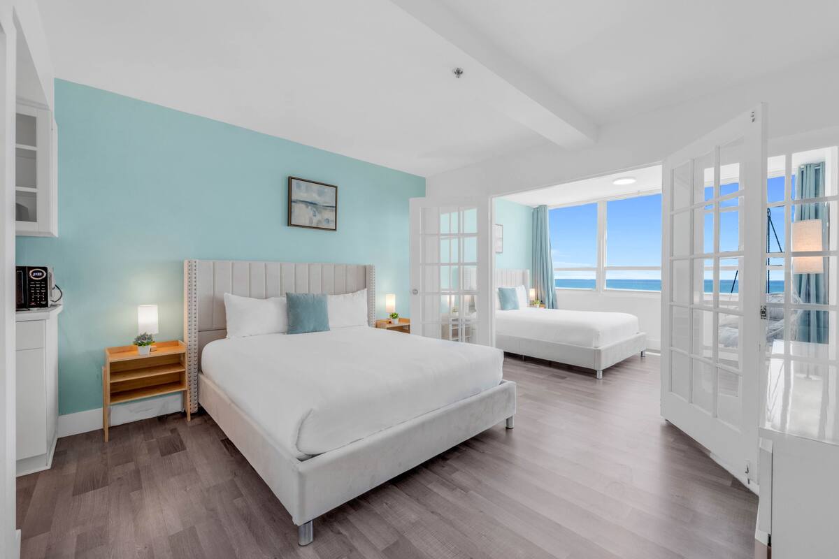 A modern hotel room with two beds, a large window with a view of the sea, and a flat-screen TV mounted on the wall.