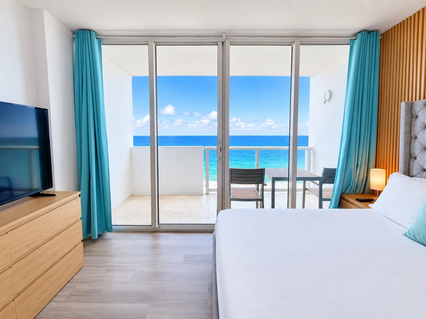 Habitación de hotel con cama, televisión, mesa y sillas, y vista al océano a través de una puerta corrediza.