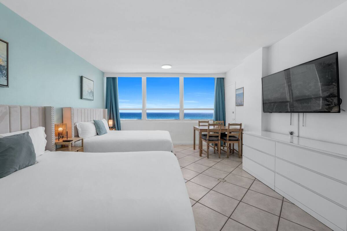 A hotel room with two beds, a TV, a desk, and a view of the sea.