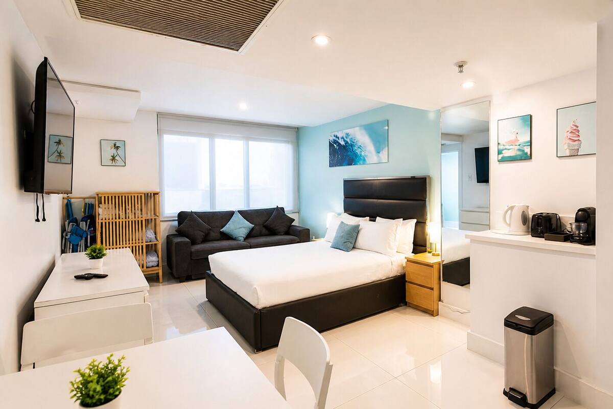 A modern hotel room with a bed, a desk, a chair, a small plant, a nightstand, a coffee machine, and artwork on the walls.