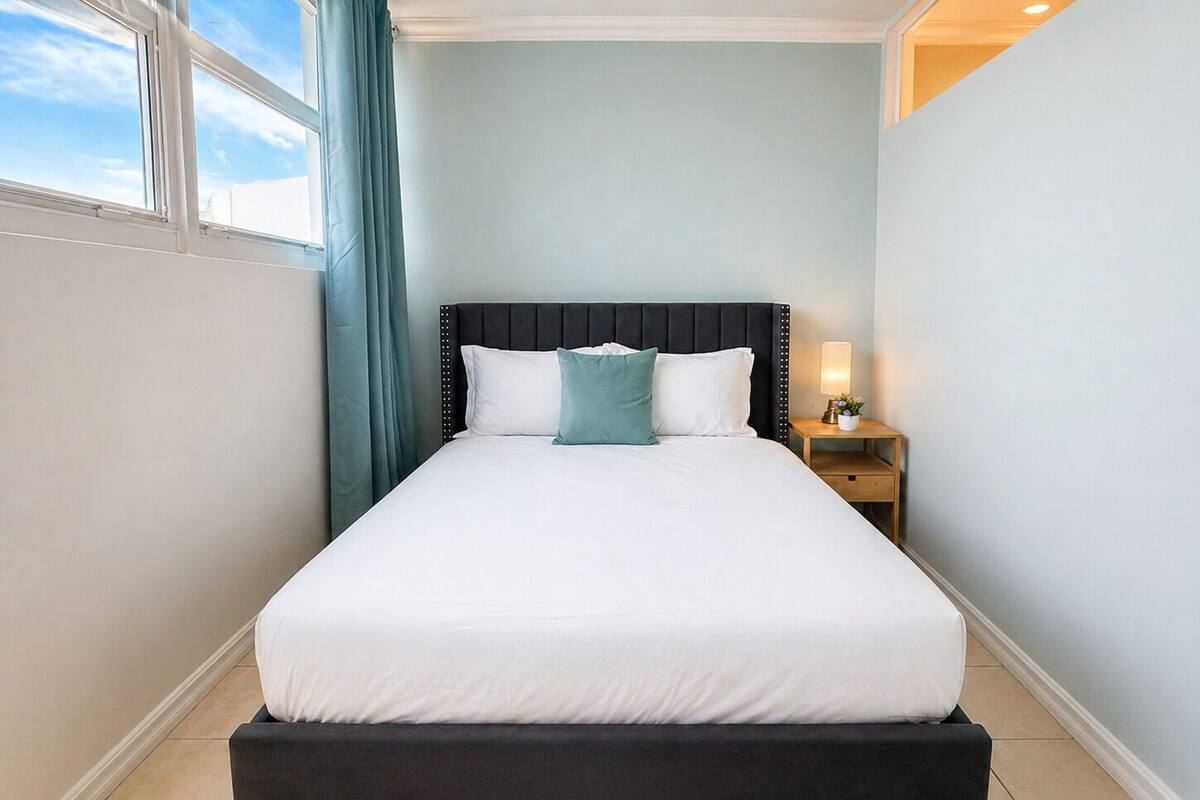 A neatly made bed with a black headboard, a bedside table with a lamp, and a window with curtains.