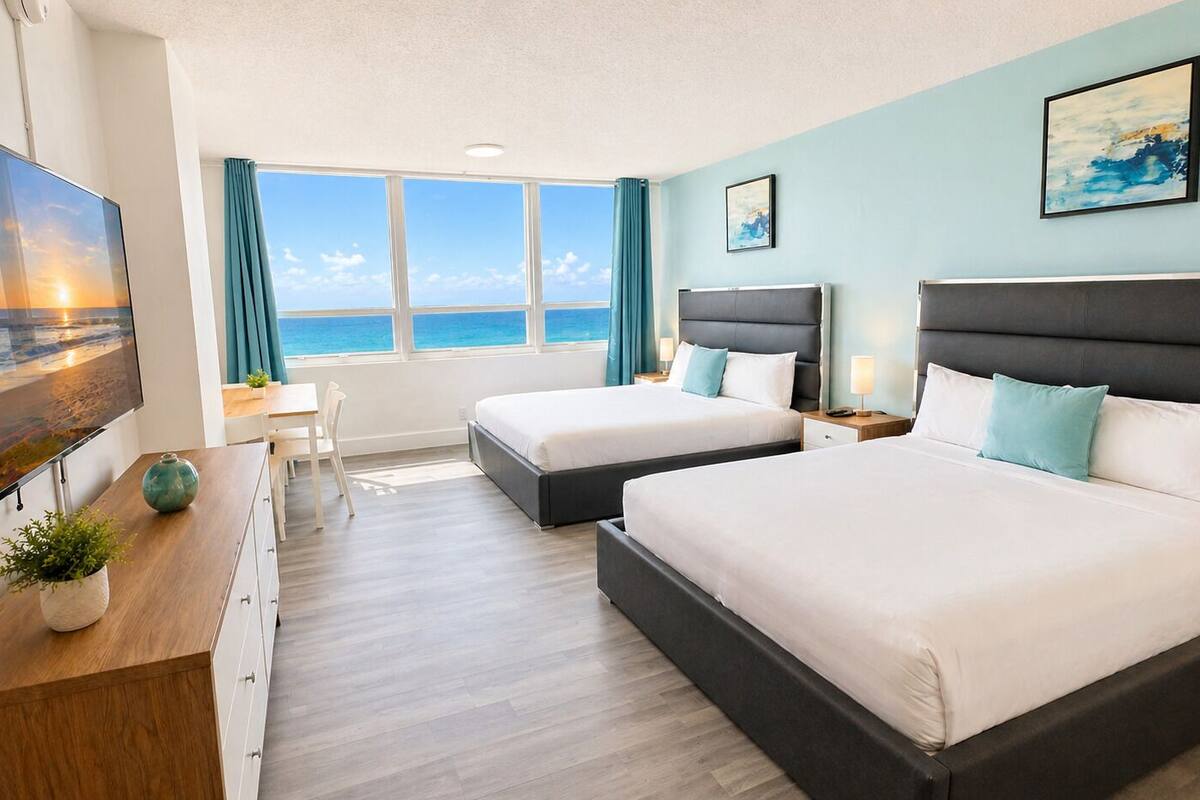 A hotel room with two beds, a desk, a TV, and a view of the ocean.
