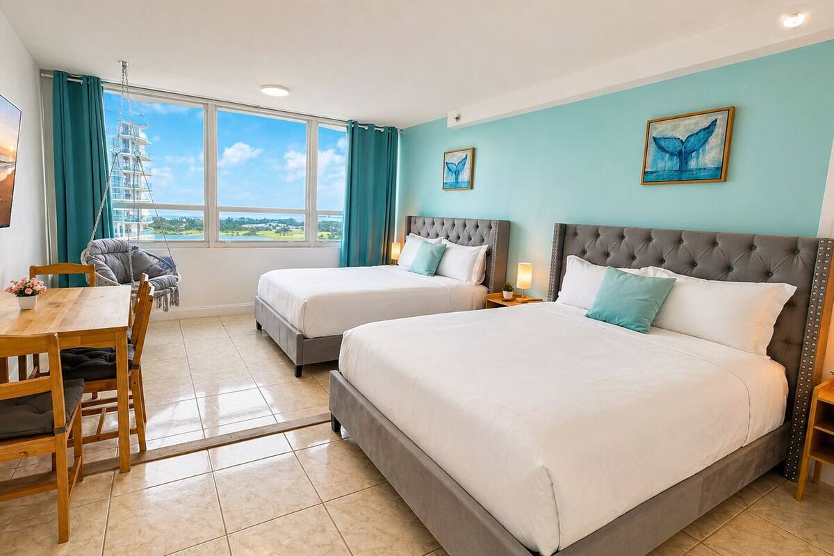 A hotel room with two beds, a dining table, chairs, a TV, and large windows with curtains.