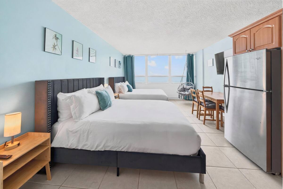 A hotel room with two beds, a dining area, a refrigerator, and a view of the sea.