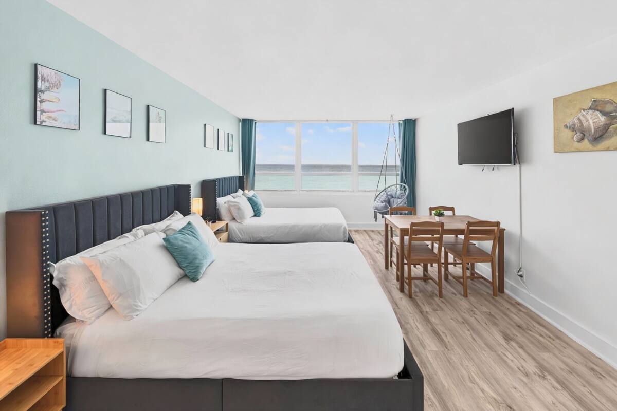 A hotel room with two beds, a TV, a dining table, and a view of the sea.