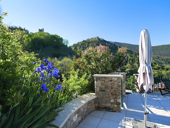 A stone patio with a white umbrella, surrounded by lush greenery and a view of a hill with a castle-like structure.