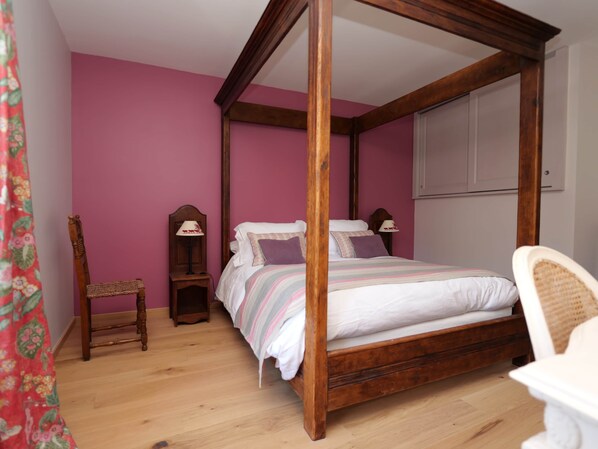 A four-poster bed with a wooden frame, a small bedside table with a lamp, a chair, and a window with a white blind.