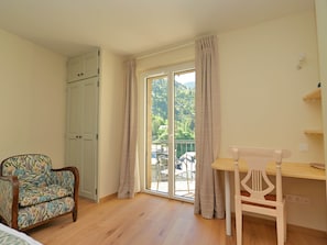 A room with a wooden floor, a desk with a chair, a patterned armchair, a sliding glass door, and a white wardrobe.