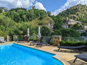 A swimming pool with lounge chairs and umbrellas, surrounded by greenery and a stone wall.