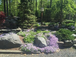 Surrounded by lush greenery, this serene garden features a birdhouse and a naturalistic landscape with large rocks and flowering shrubs.