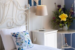 Un lit bien fait, agrémenté d’un coussin à motifs floraux, une table de chevet avec une lampe et une petite table sur laquelle est posé un vase de fleurs.