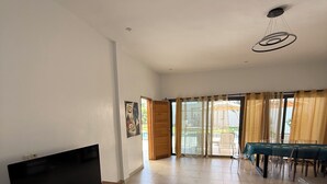 Amplia sala de estar con una televisión de pantalla plana sobre un mueble de madera, comedor con mesa y sillas, y puerta corrediza de vidrio que da a un patio al aire libre.
