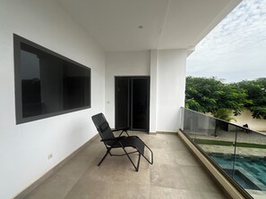 A balcony with a black chair, a large window, and a view of greenery.