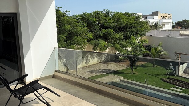Un balcón con dos sillones negros, vista despejada de un césped verde y árboles, y un edificio blanco al fondo.