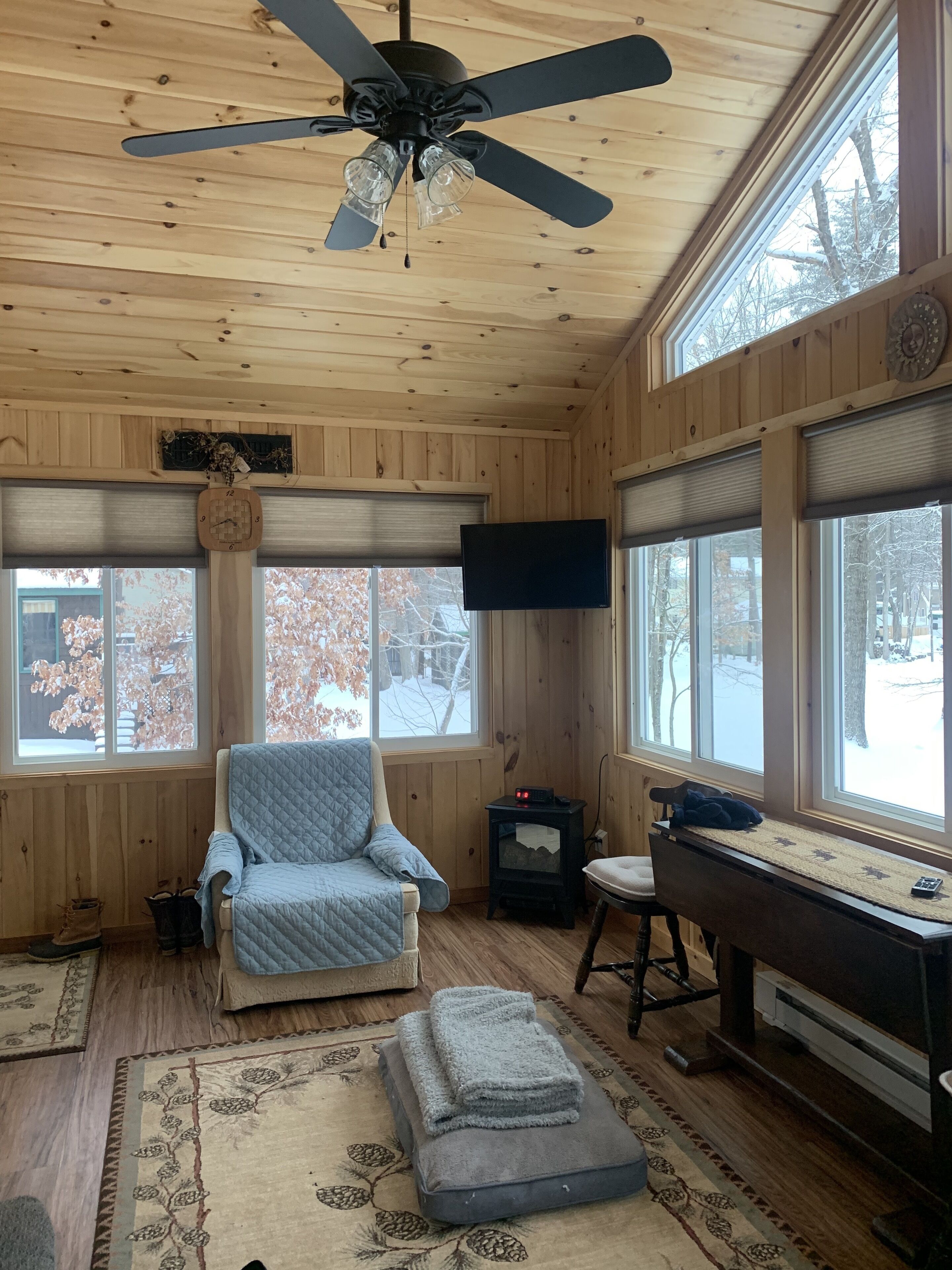 Interior acogedor de una cabaña con ventilador de techo, un sillón acolchado, un escritorio con computadora y ventanas amplias que ofrecen vista a un paisaje nevado.