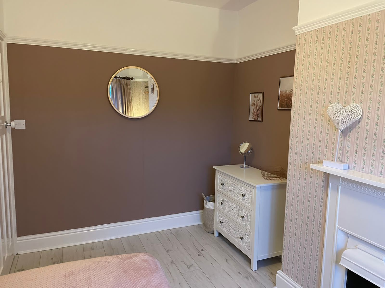 Ein Schlafzimmer mit brauner Wand, einem weißen Schminktisch, einem runden Spiegel, einem rosa Bett und einem Kamin.