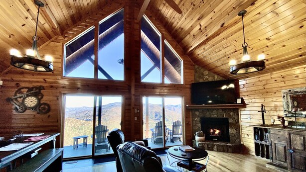 Una cabaña espaciosa con techo alto, ventanas grandes con vistas a las montañas, chimenea y mobiliario de madera.
