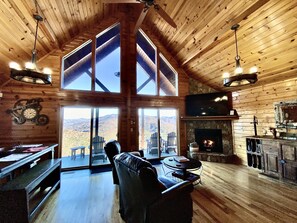 A spacious cabin with a high ceiling, large windows offering mountain views, a fireplace, and wooden furnishings.
