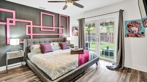 Ein modernes Schlafzimmer mit einem großen Bett, einem Deckenventilator, einem Nachttisch und Blick durch eine Glastür in einen Außenbereich.