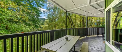 Una terraza techada al aire libre con una mesa de picnic y bancos, rodeada de abundante vegetación y árboles.