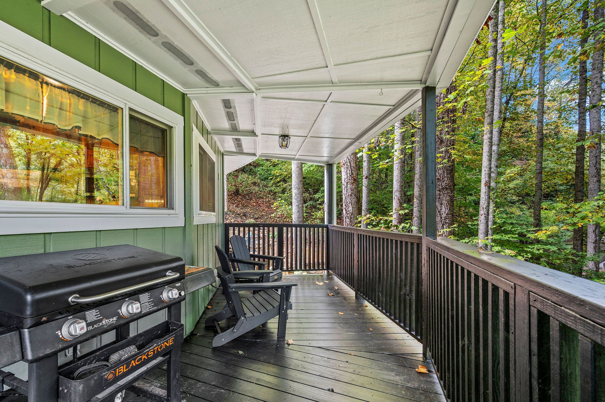 A covered porch with a grill, chairs, and a view of a forest.