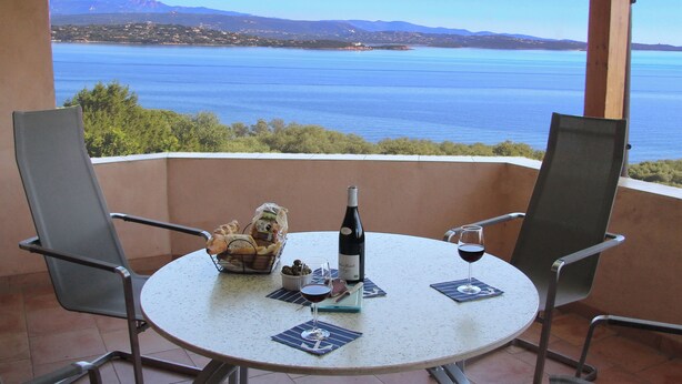A round table with two chairs, a bottle of wine, a glass of wine, and a basket of bread and olives on a balcony overlooking a body of water with mountains in the background. - Three-flag house with a view of the Gulf of Porto-Vecchio / quiet location (Porto-Vecchio)
