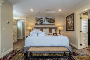 Habitación de hotel con una cama grande, dos lámparas de noche, un cuadro de barcos y una puerta a la derecha.