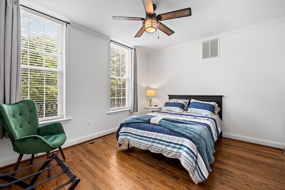 A bedroom with a bed, a chair, a ceiling fan, and two windows.