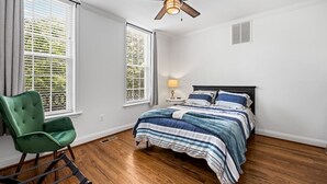 A bedroom with a bed, a chair, a ceiling fan, and two windows.
