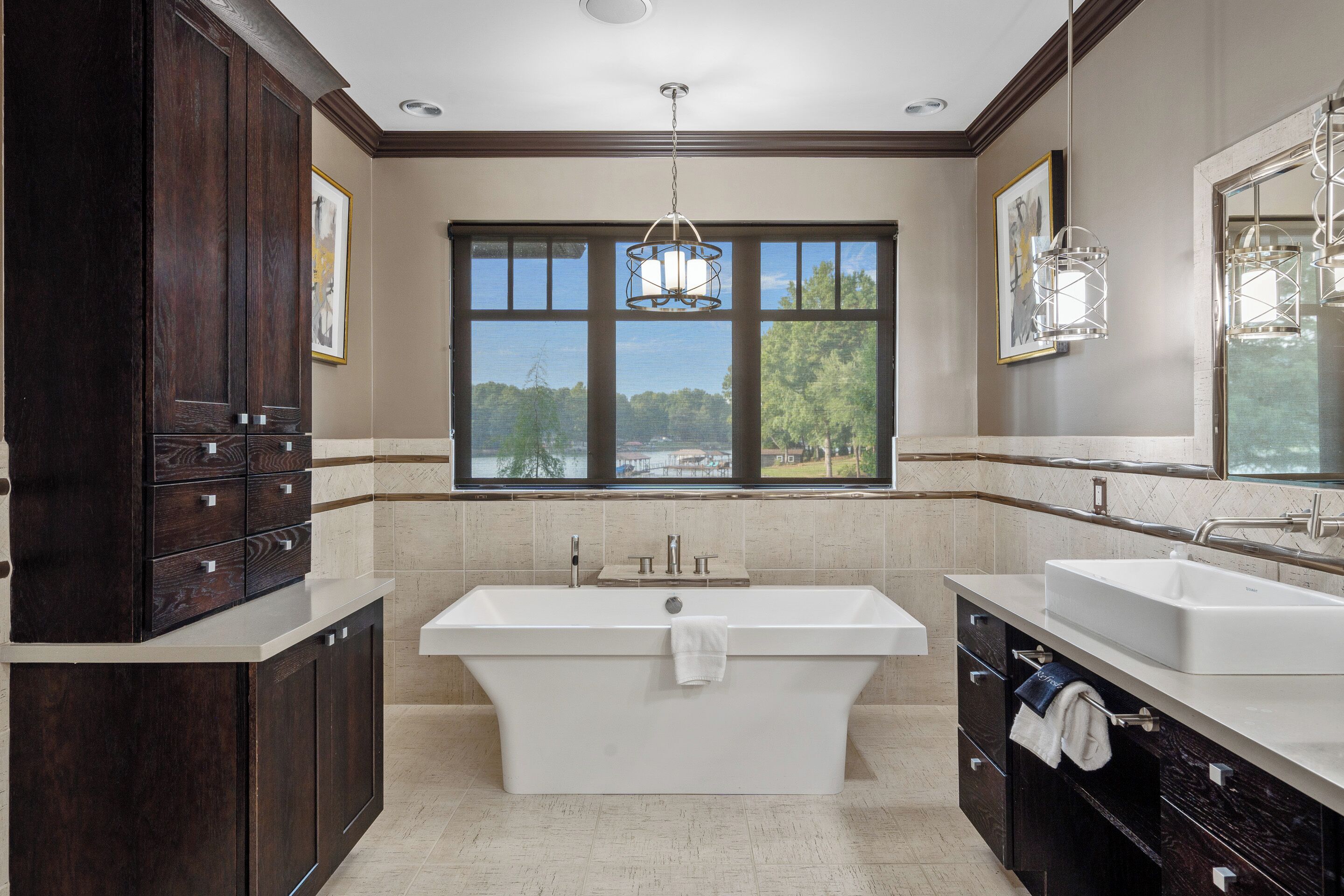 Un baño moderno con una bañera independiente, lavamanos dobles, muebles de madera oscura y un amplio ventanal con vistas a árboles.
