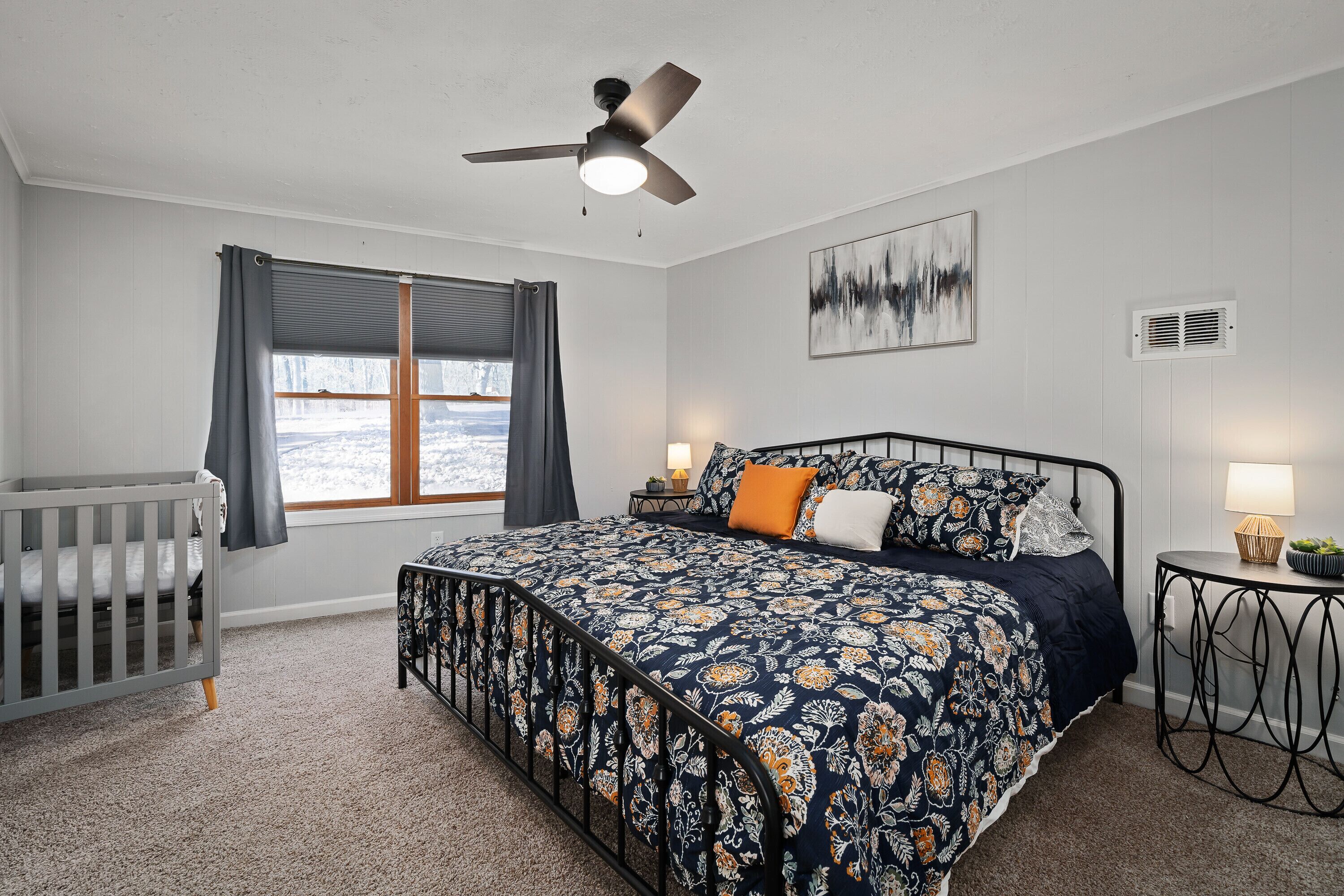 Une chambre à coucher comprenant un lit, un berceau, un ventilateur de plafond, une fenêtre avec des rideaux, une table de chevet avec une lampe et une bouche d’aération fixée au mur.