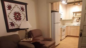 Una sala de estar con ventilador de techo, lámpara y sofá. Se ve una cocina a través de un vano abierto.