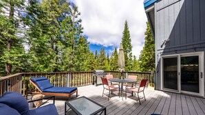 A wooden deck with outdoor furniture, including lounge chairs and a table, surrounded by trees. - Cheerful, spacious family cabin ready for fun! (Shaver Lake)