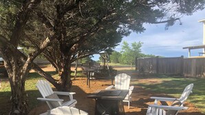 An outdoor seating area with Adirondack chairs and a table, surrounded by trees and a wooden fence. - Large Open Range Camper sleeps up to 4 people (Dripping Springs)