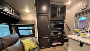 A compact RV interior with a leather sofa, a built-in entertainment unit, a kitchenette, and a window with a view. - Large Open Range Camper sleeps up to 4 people (Dripping Springs)