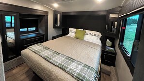 A bedroom with a bed, a TV, a window, and a headboard. - Large Open Range Camper sleeps up to 4 people (Dripping Springs)