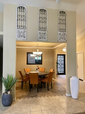 A dining area with a round table, six chairs, a chandelier, and decorative ironwork above the windows.