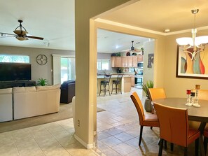 A modern living room with a sofa, TV, and ceiling fan. A dining area with a table and chairs is visible in the background.
