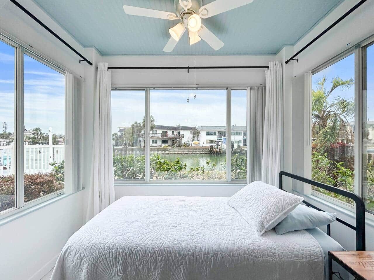 Un dormitorio con cama, ventilador de techo, ventanas amplias y vistas a edificios y zonas verdes.