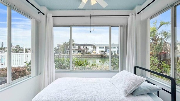 Ein Schlafzimmer mit Bett, Deckenventilator, großen Fenstern und Blick auf Gebäude und Grünflächen.