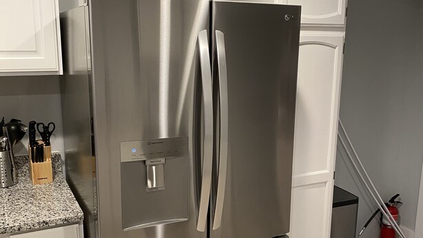 Un refrigerador de acero inoxidable con dispensador de agua, situado en una cocina con gabinetes blancos y encimeras de granito.