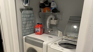 Zona de lavandería con lavadora y secadora de carga frontal, repisa con productos de limpieza y un perchero con una prenda a rayas.